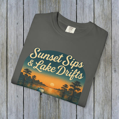Folded Lake Drifts T-Shirt in soft grey, featuring the captivating 'Sunset Sips & Lake Drifts' graphic on a wooden backdrop.