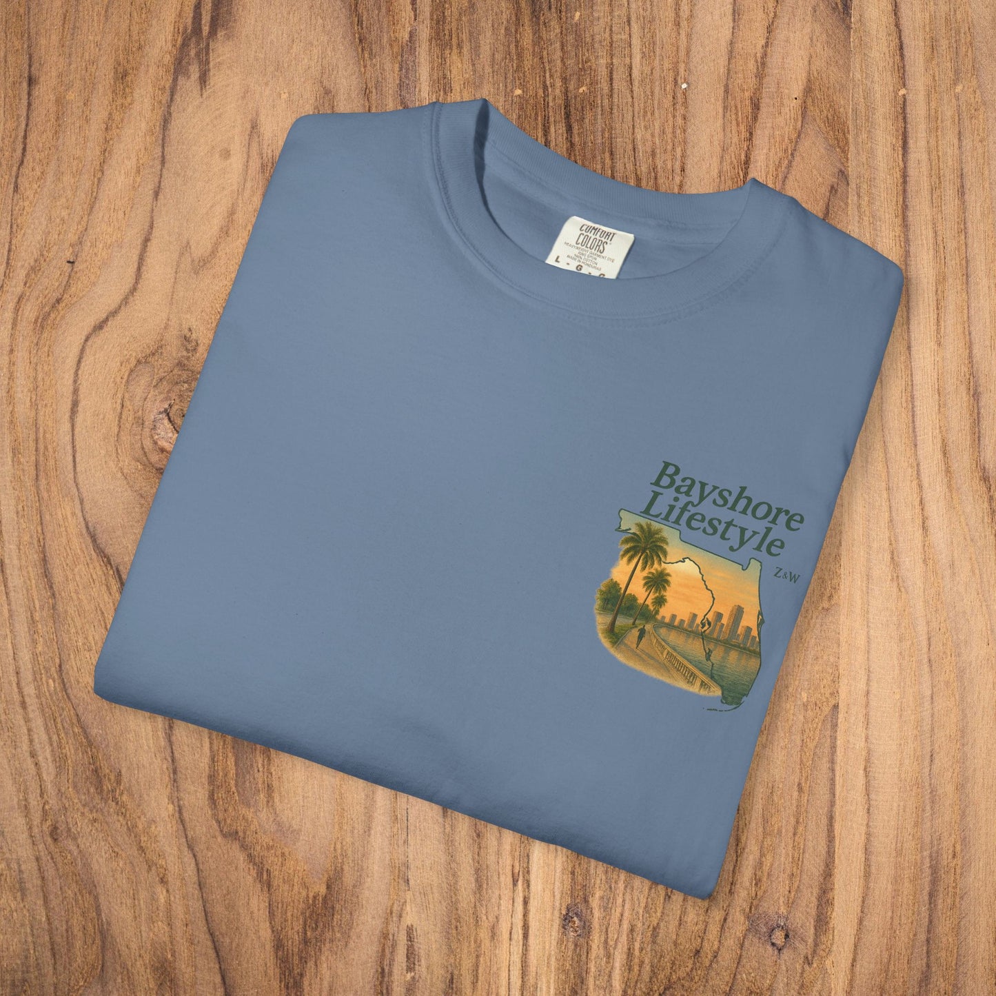 Bayshore Lifestyle Tee - 100% Cotton Lightweight Fabric, Ribbed Neck, Sizes XS-5XL - Folded blue t-shirt with graphic design on wood background.