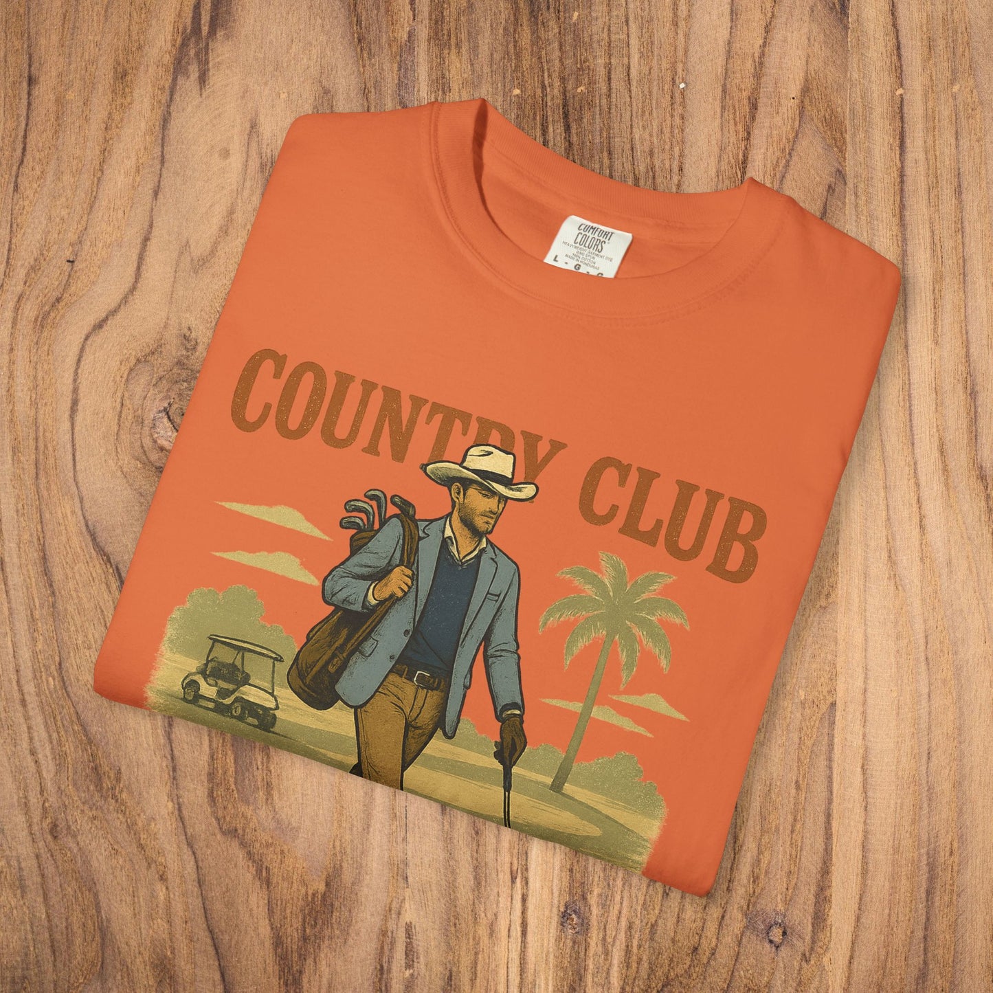 Country Club Cowboy Graphic Tee in an orange color, beautifully arranged on a wooden surface, emphasizing its cowboy graphic design.