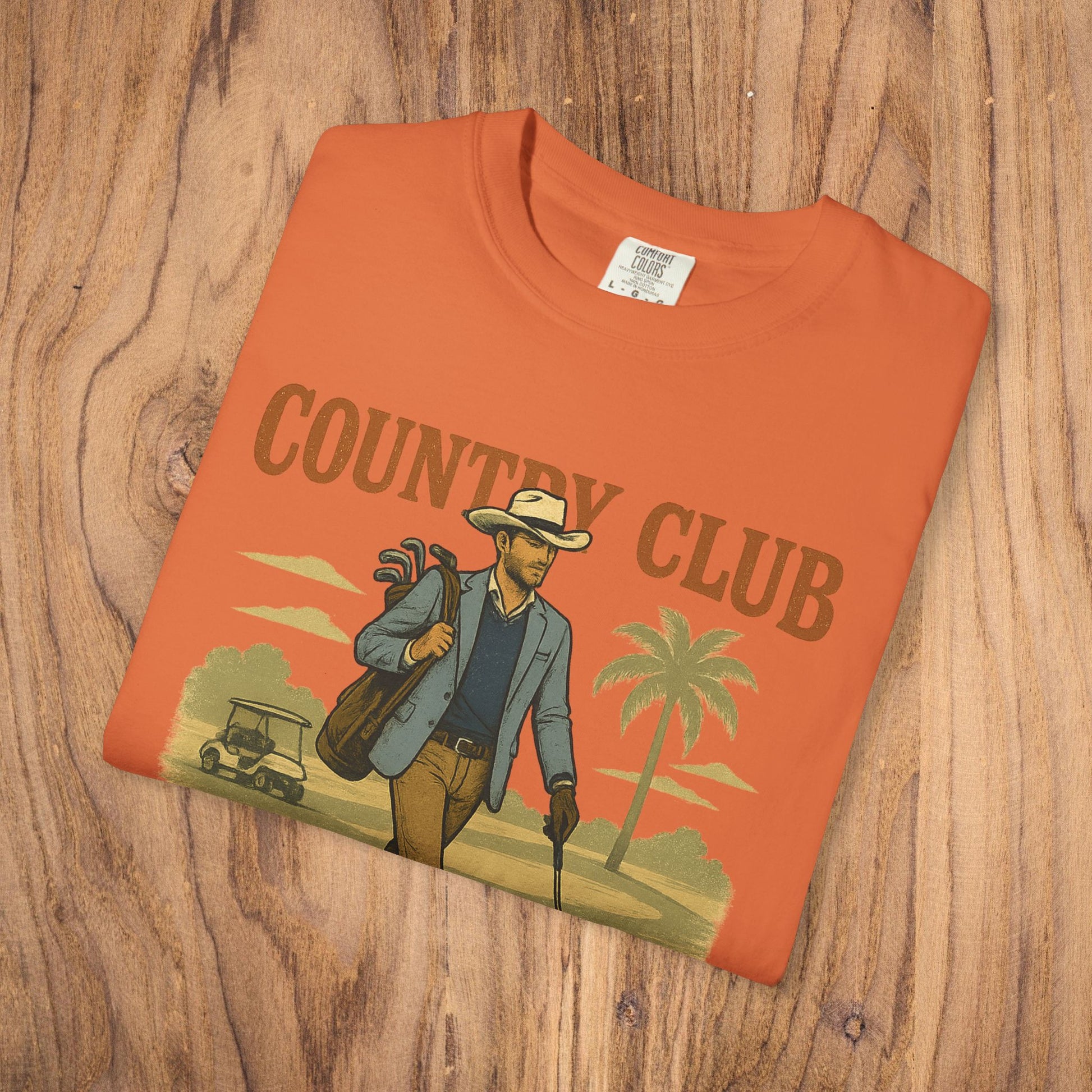Country Club Cowboy Graphic Tee in an orange color, beautifully arranged on a wooden surface, emphasizing its cowboy graphic design.
