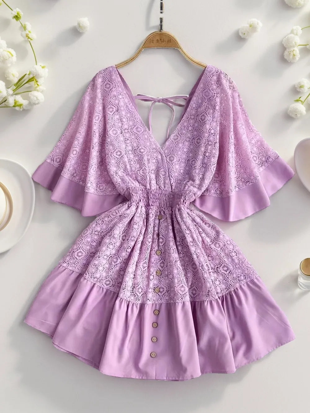Flat lay image of a Semi-Sheer Ruffle Hem Lace Mini Dress with Tie Back - Elegant Smocked Design in a soft lavender shade, featuring ruffles and button accents, perfect for warm weather wear.