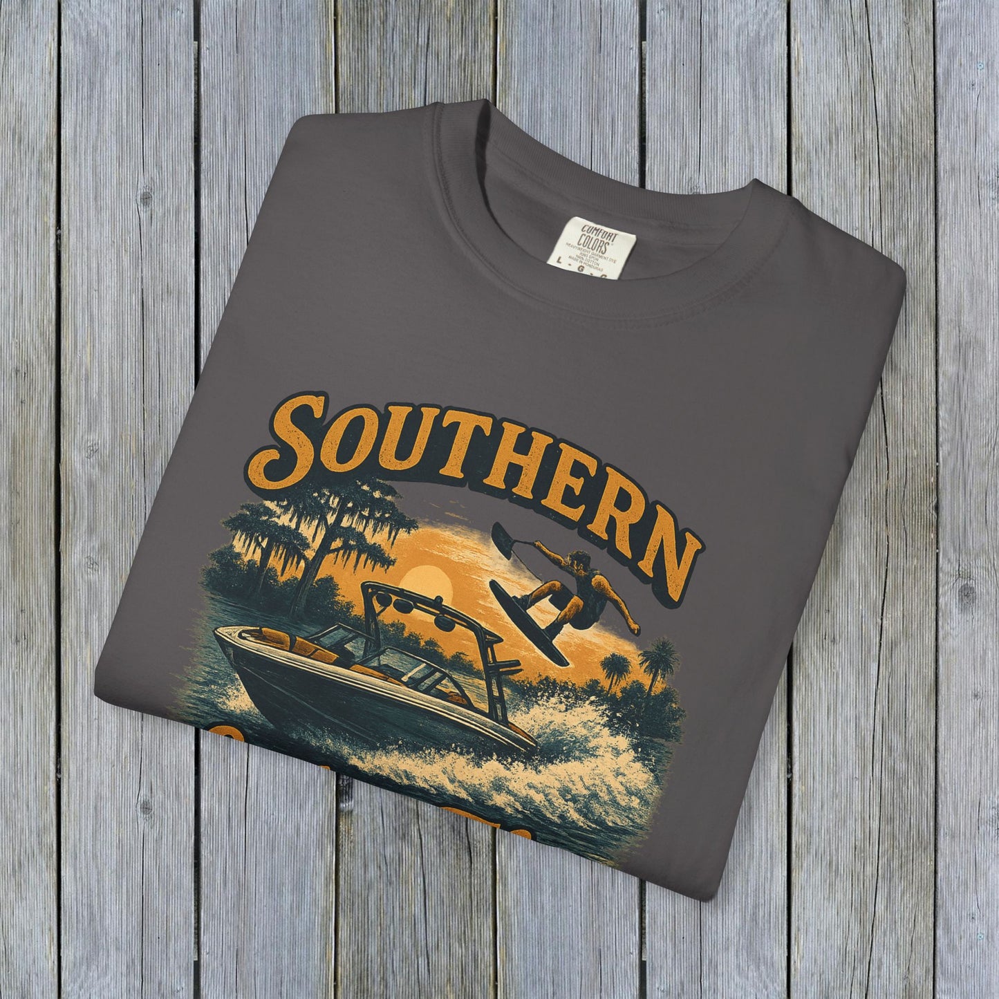 Southern Lake Flow T-Shirt laid flat on a wooden backdrop, in grey color with a graphic of a boat and wakeboarder, emphasizing durability and sustainable materials from 100% US cotton.