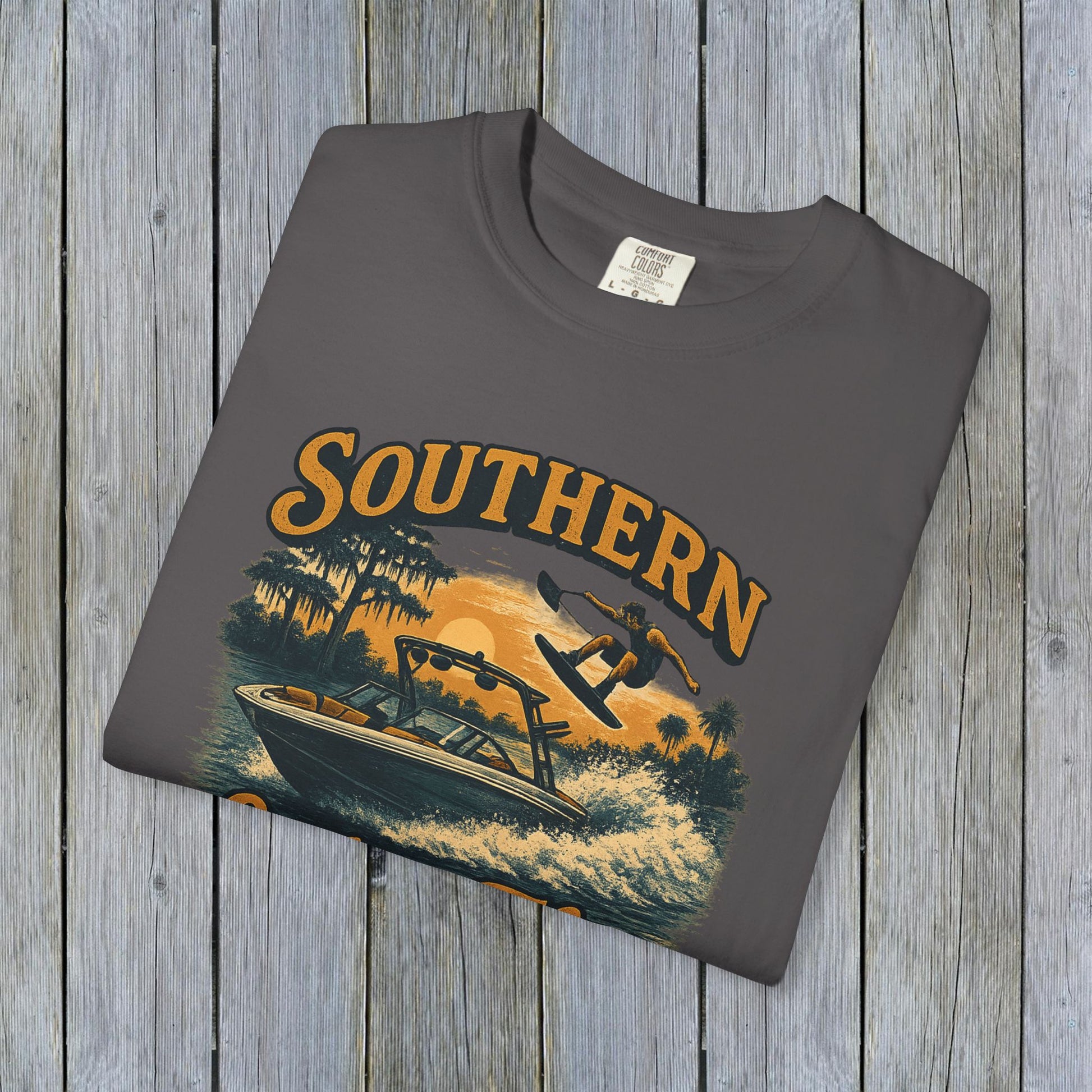 Southern Lake Flow T-Shirt laid flat on a wooden backdrop, in grey color with a graphic of a boat and wakeboarder, emphasizing durability and sustainable materials from 100% US cotton.