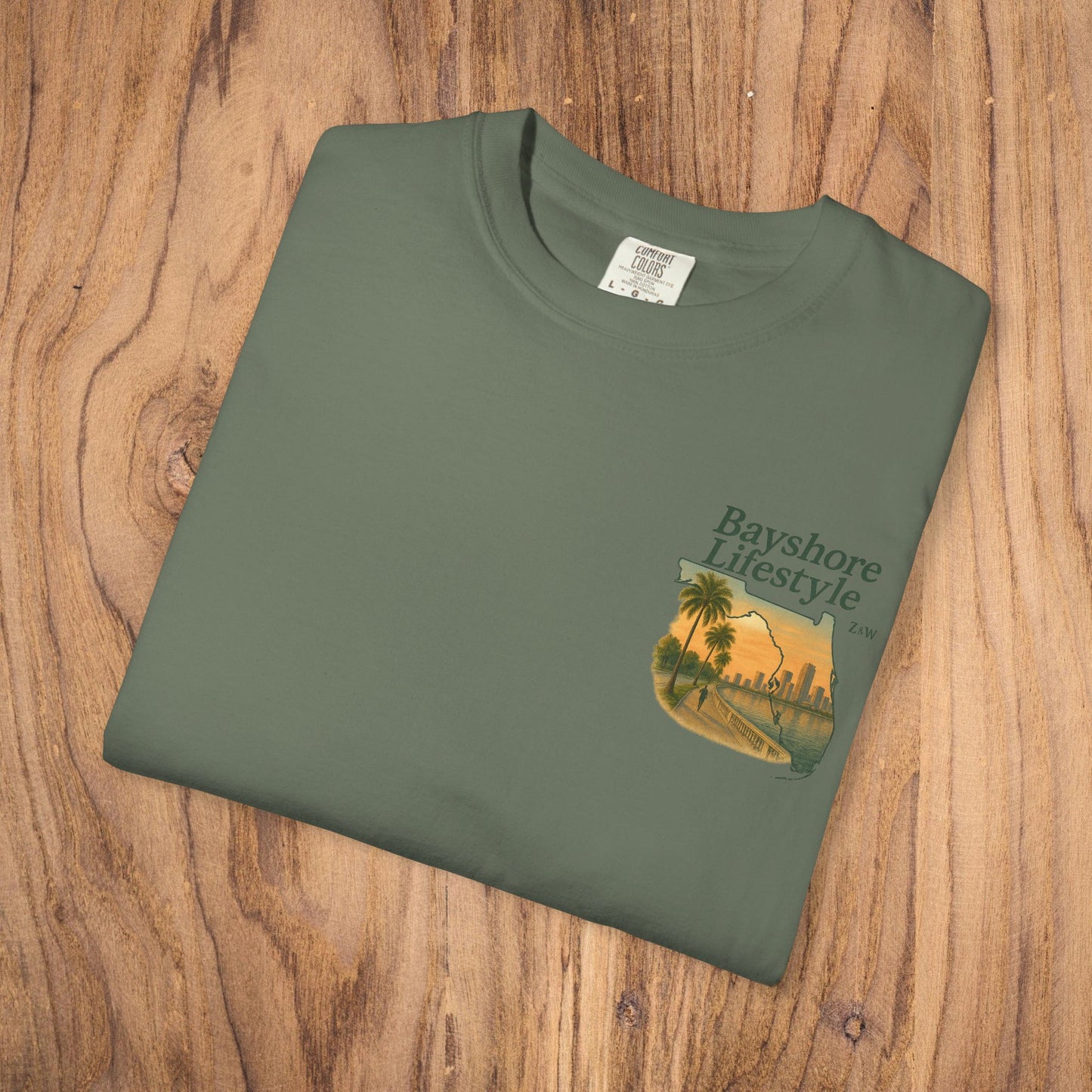 Bayshore Lifestyle Tee - 100% Cotton Lightweight Fabric, Ribbed Neck, Sizes XS-5XL - Folded green t-shirt featuring a graphic design on wood background.