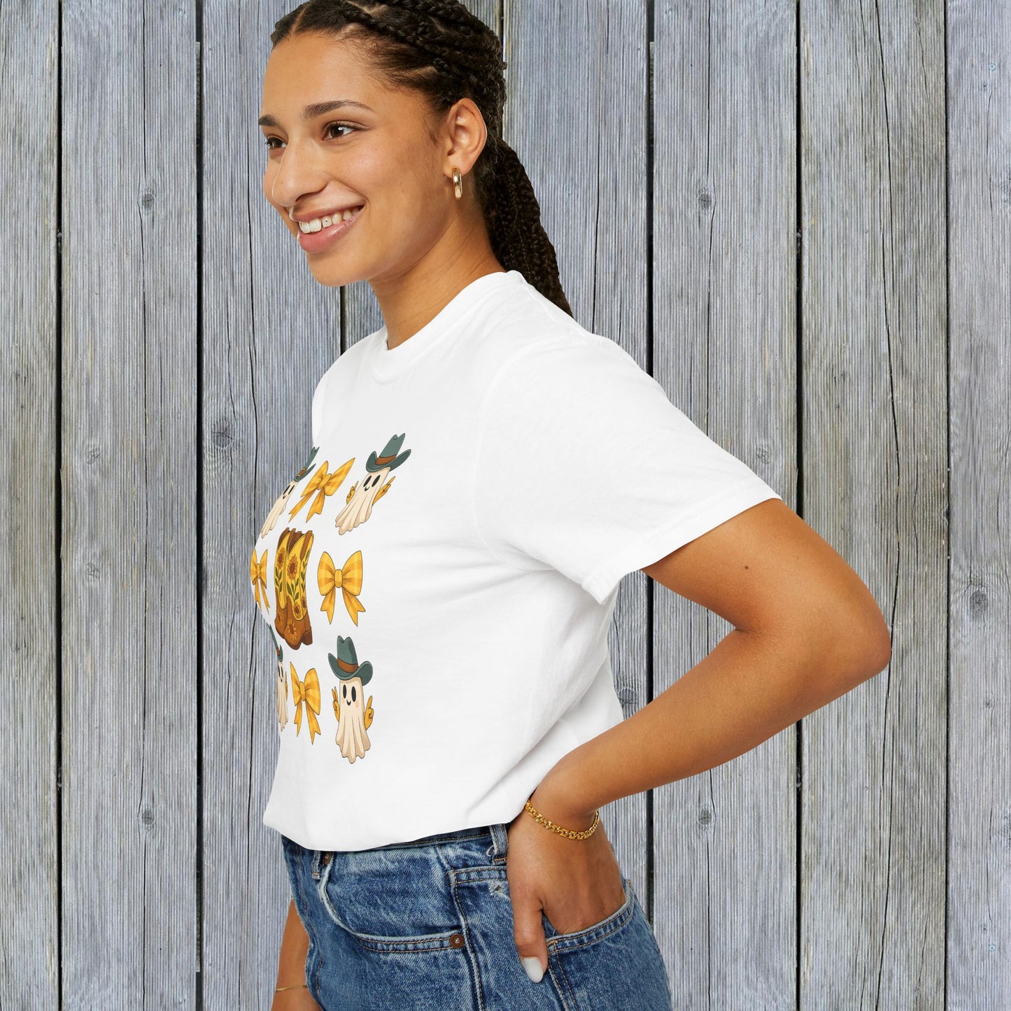 Cute Halloween T-Shirt worn by a smiling woman, illustrating fun ghosts and cowboy boots, showcased against a textured wooden wall, great for unisex fall fashion lovers.