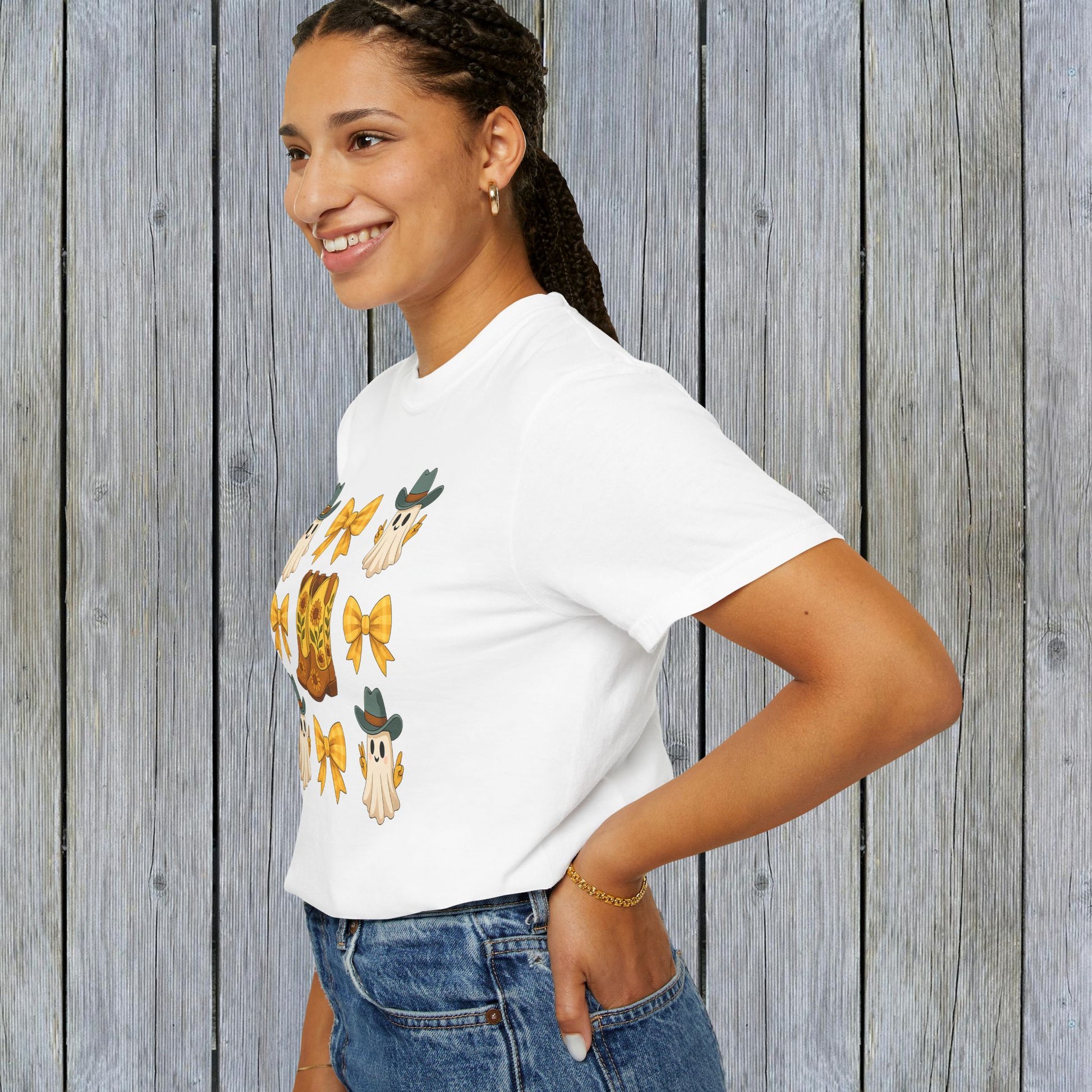 Cute Halloween T-Shirt worn by a smiling woman, illustrating fun ghosts and cowboy boots, showcased against a textured wooden wall, great for unisex fall fashion lovers.
