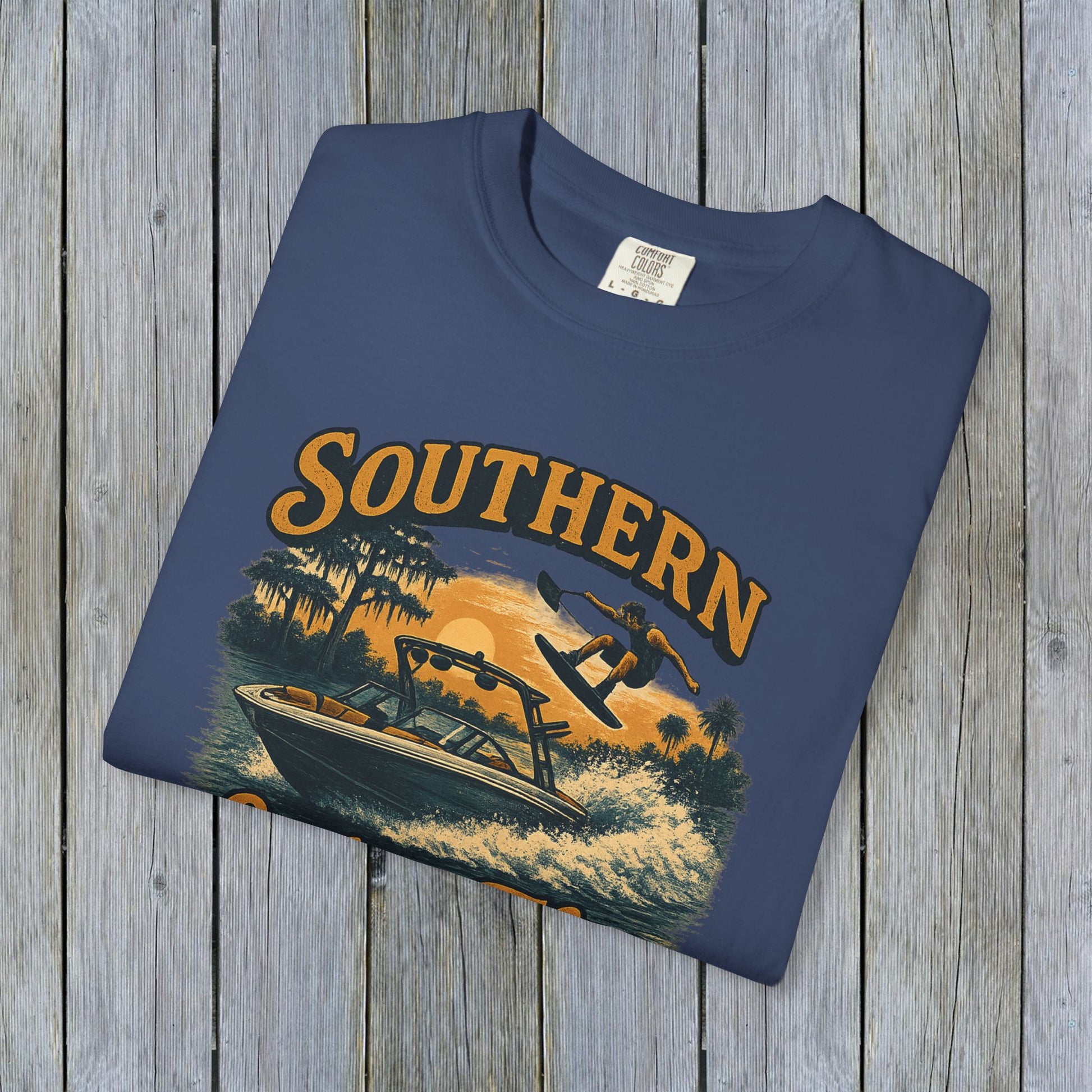 Southern Lake Flow T-Shirt displayed on a wooden surface, showcasing eco-friendly design with a graphic featuring a boat and wakeboarder, crafted from 100% US cotton for a stylish and durable fit.