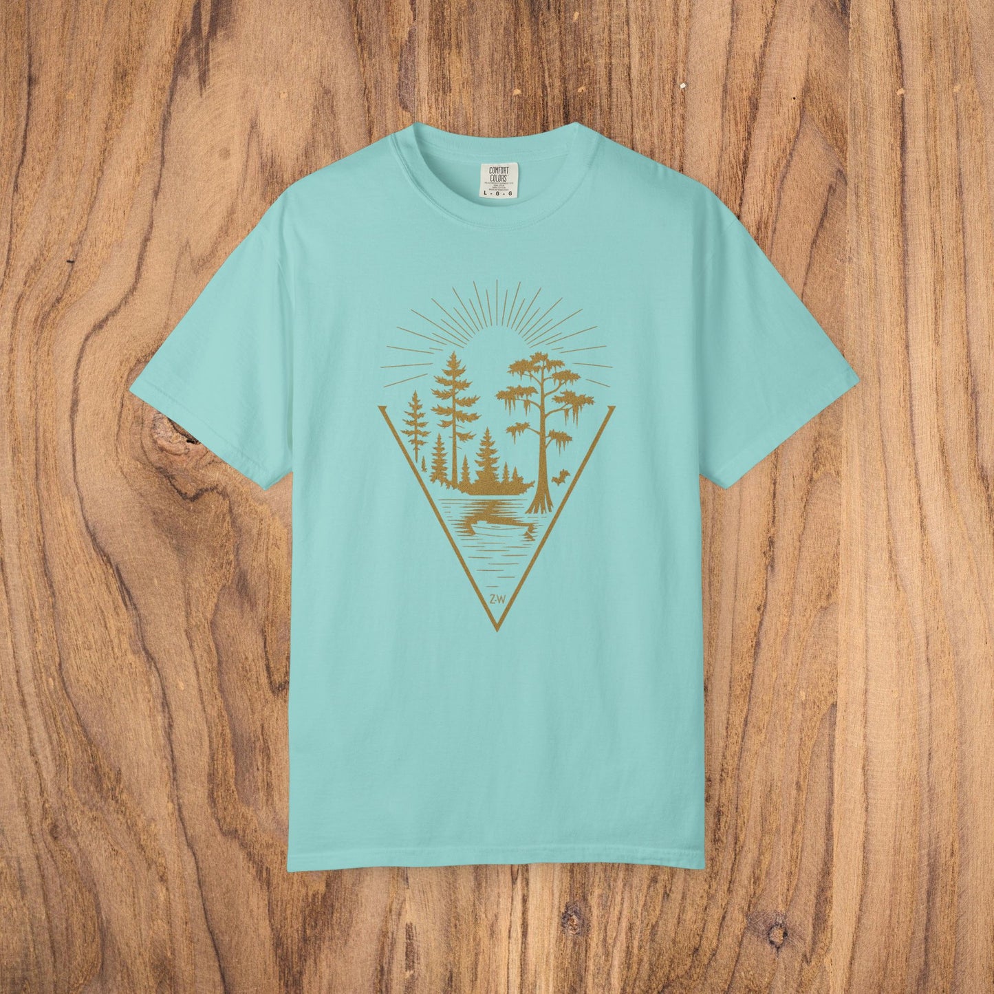 Nature Outdoor Vibes Tee displayed on a wooden background, showing the unique graphic design of trees and a lake in gold on a light teal shirt, crafted for comfort from 100% US cotton.