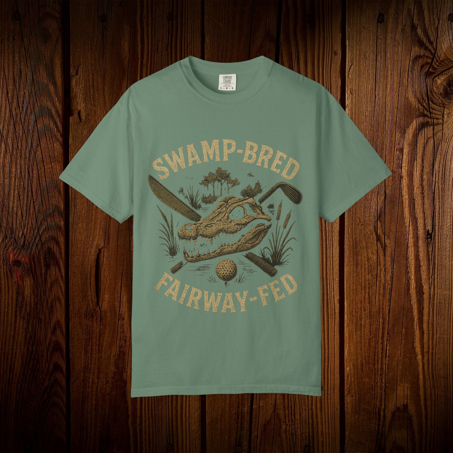 Unisex Swamp-Bred Fairway-Fed Tee in soft green, highlighting a striking graphic of an alligator skull with crossed golf clubs, photographed against a rustic wooden setting.