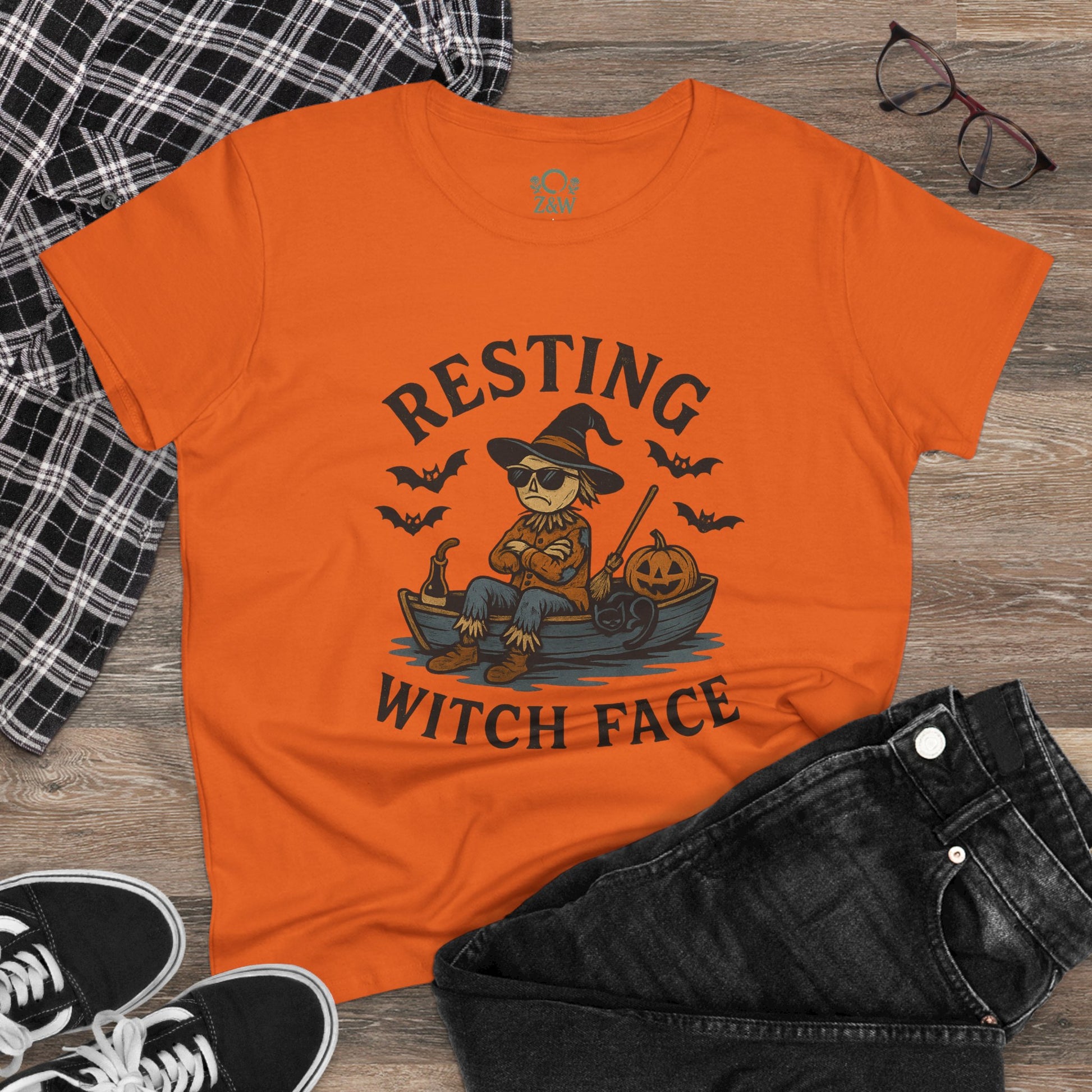Casual flat lay of the Resting Witch Face Halloween T-Shirt for Women in vivid orange with stylish black jeans, showcasing Halloween spirit and humor, ideal for festive occasions.