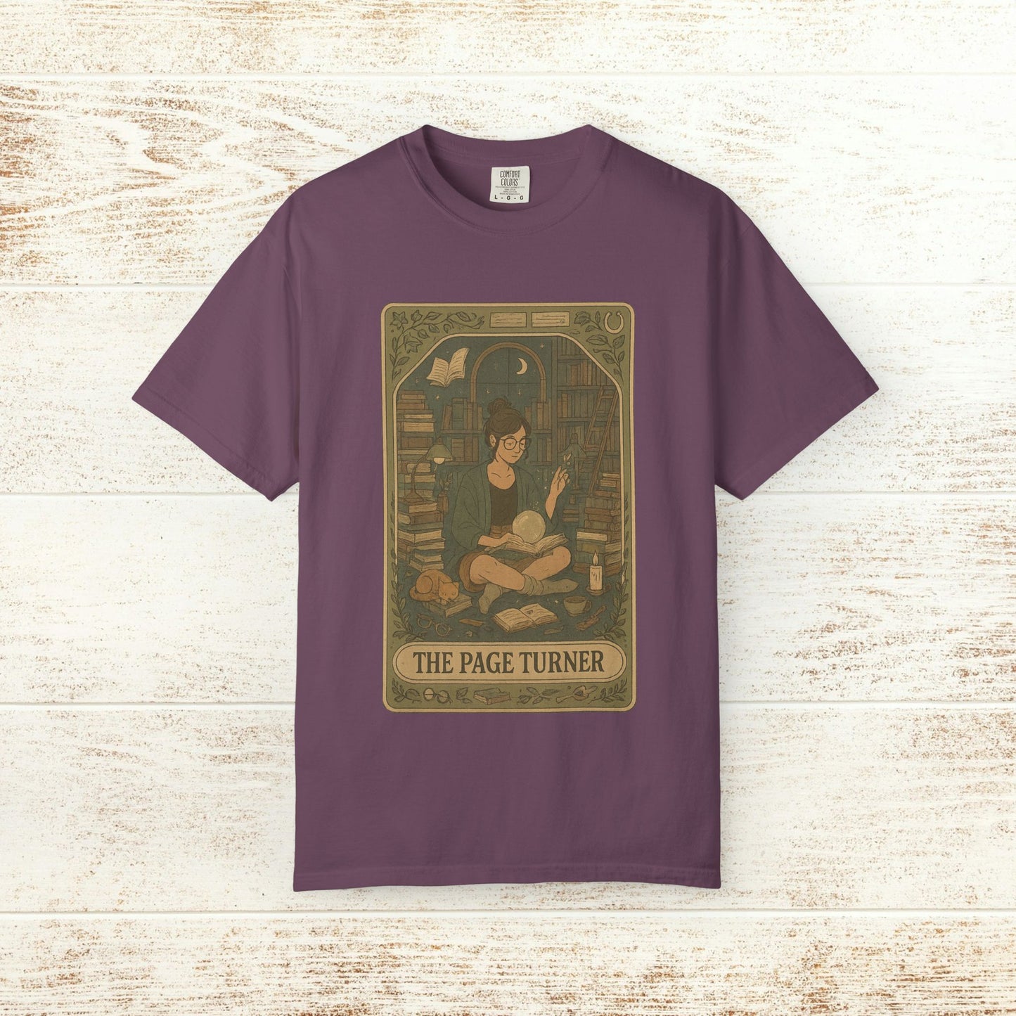 Page Turner Tarot Tee - A stylish purple soft cotton t-shirt displaying an enchanting illustration of a reader in a cozy library, available in sizes S to 4XL.