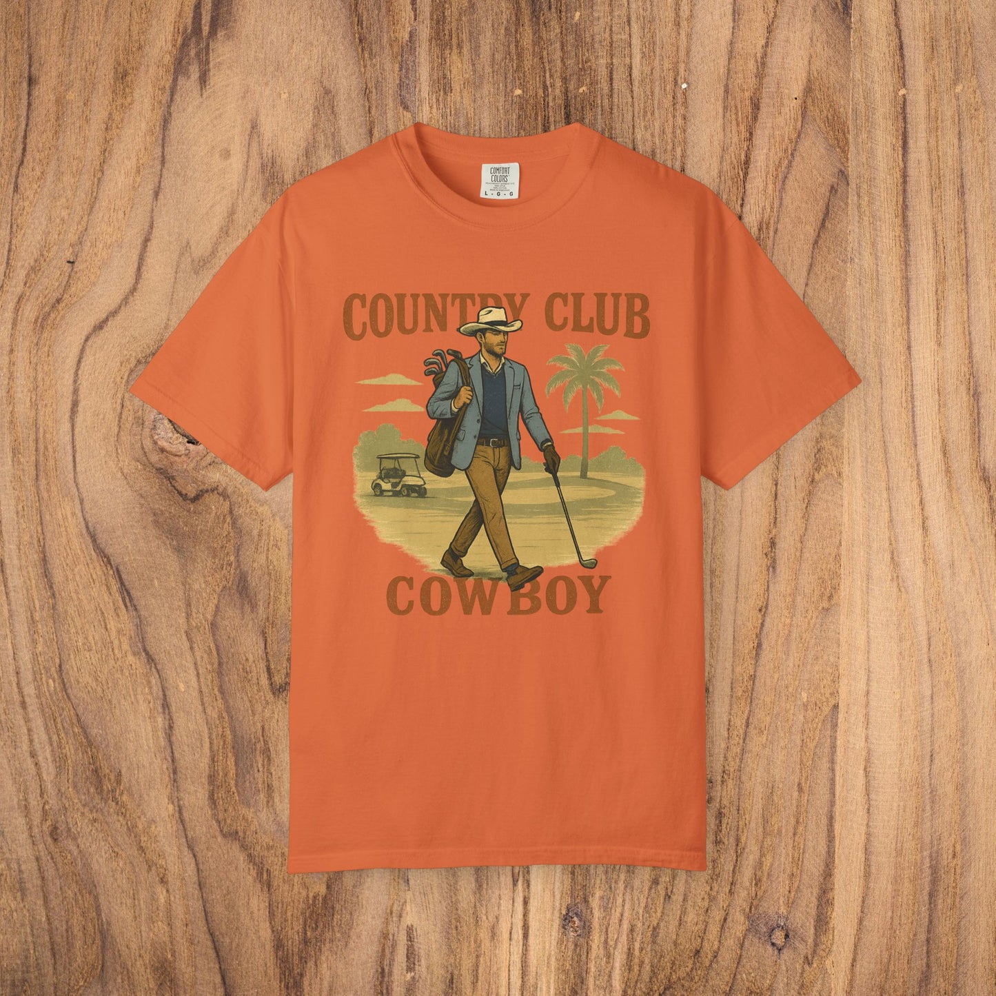 Country Club Cowboy Graphic Tee in vibrant orange, highlighting a cowboy with golf clubs in a casual design. Constructed from durable cotton fabric, perfect for outdoor activities.
