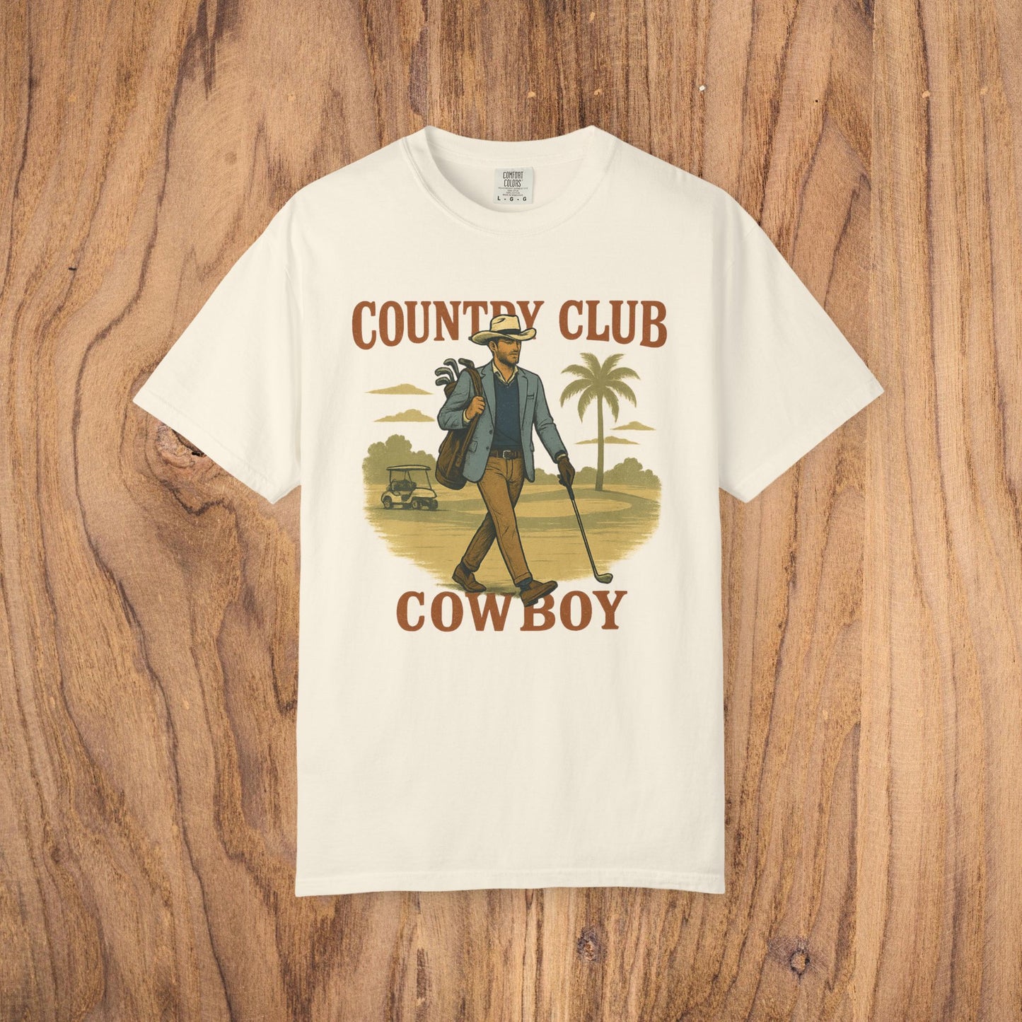 Country Club Cowboy Graphic Tee in cream color, featuring a cowboy holding golf clubs against a tropical background. Made from durable cotton fabric with a ribbed collar for added comfort.