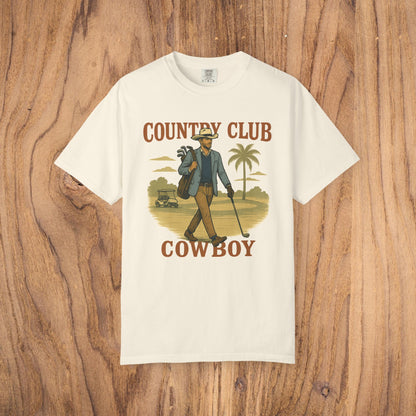 Country Club Cowboy Graphic Tee in cream color, featuring a cowboy holding golf clubs against a tropical background. Made from durable cotton fabric with a ribbed collar for added comfort.