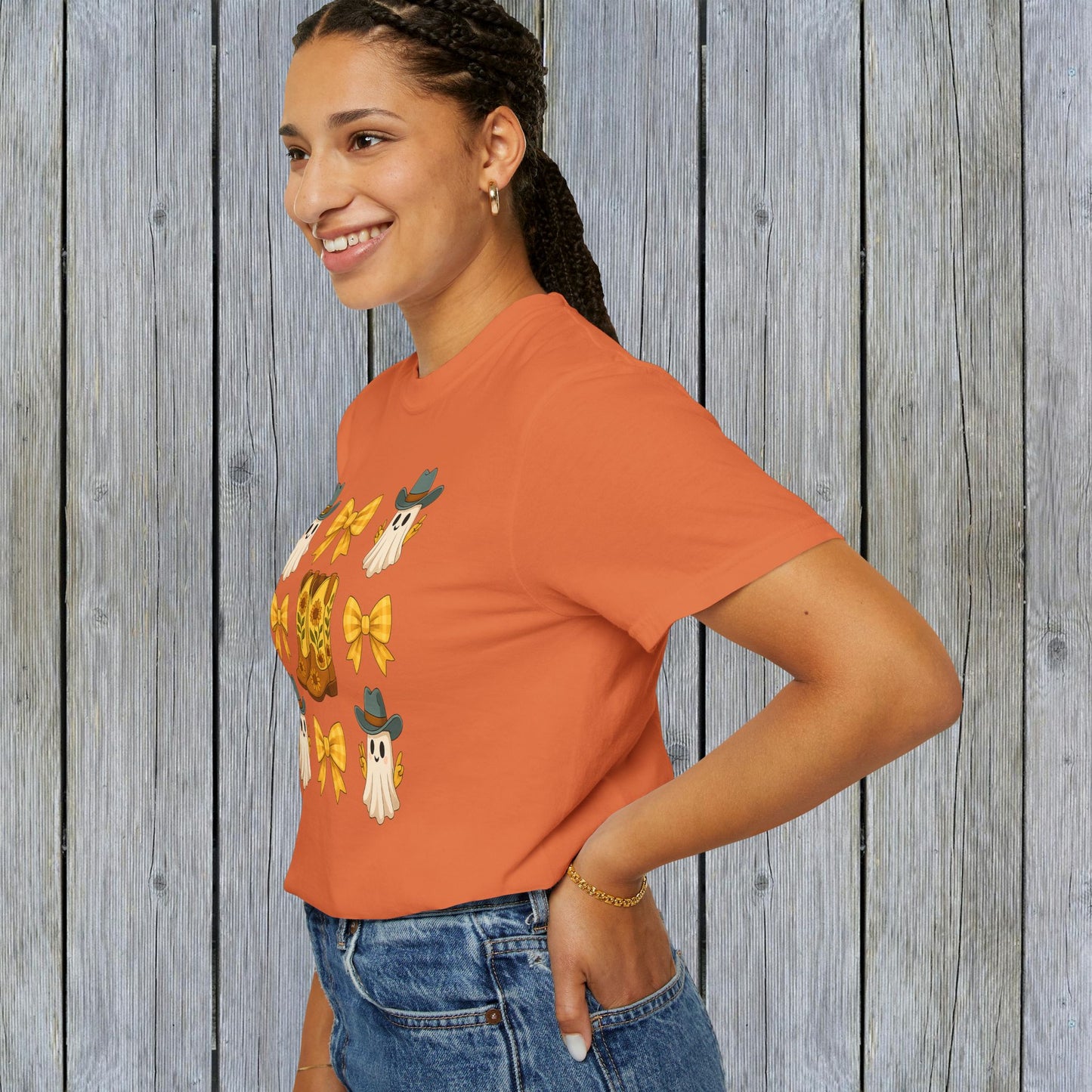 Cute Halloween T-Shirt featuring ghosts and cowboy boots, shown worn by a woman smiling in a side profile against a rustic wooden background, part of unisex fall fashion collection, made from eco-friendly fabric.