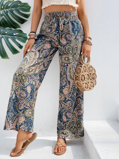 Model wears Trendy Printed Wide Leg Pants with Tied Detail, beautifully paired with a woven bag, highlighting the elegant paisley print made from 100% opaque polyester.