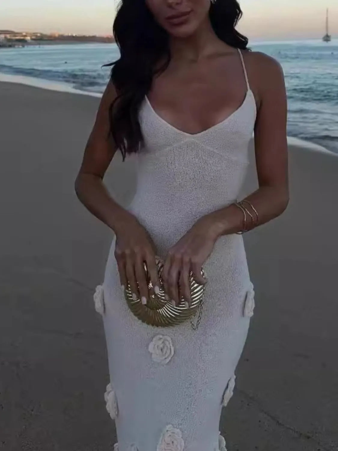 Close-up of a model wearing a backless flower print maxi sweater cami dress on the beach, holding a chic gold clutch. This slightly stretchy 100% polyester dress is ideal for summer elegance.